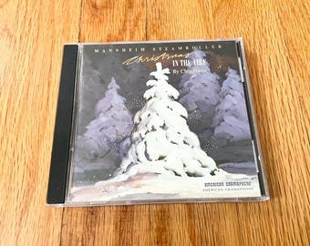 May include: A CD case featuring the album "Christmas in the Aire" by Mannheim Steamroller. The cover art depicts a snowy Christmas tree and other snow-covered trees against a gray background. The text "Christmas in the Aire" is written in a stylized font.