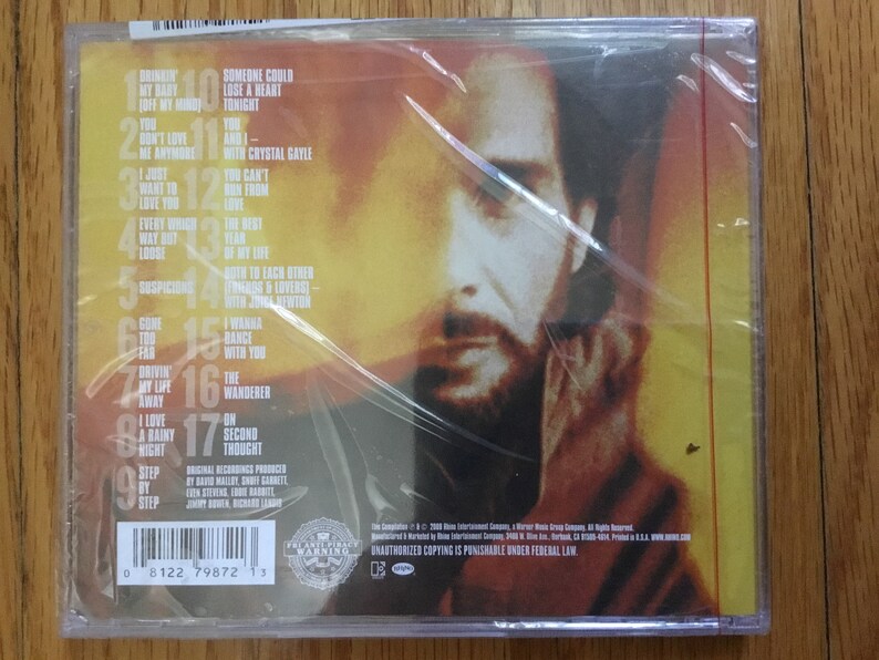 May include: The back of a CD case with a close-up of a man's face. The case is yellow and red with a list of song titles and track numbers. The text on the case reads "Unauthorized copying is punishable under federal law."