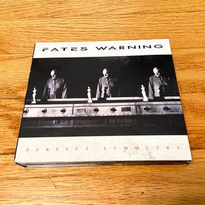 May include: A square CD case featuring the Fates Warning album Perfect Symmetry. The cover art displays three identical figures in dark shirts standing behind a metallic control panel with small white statues, set against a black background.
