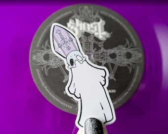 May include: A white sticker featuring a cartoon ghost wearing a bishop's mitre with a cross design. The sticker is placed on a dark gray circular coaster with the band name "Ghost" printed on it. The background is a vibrant purple.