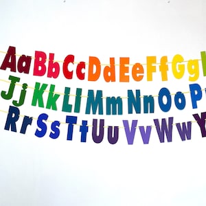 Może przedstawiać: Baner alfabetu tęczy z literami wyciętymi z kolorowego papieru i nawleczonymi na żółtą sznurówkę. Litery są wielkimi literami i są ułożone alfabetycznie.