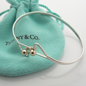 May include: A silver bangle bracelet with a gold accent and two small gold balls. The bracelet is on a turquoise blue velvet pouch with the text "Tiffany & Co."