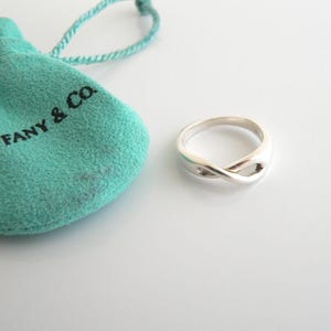 May include: A silver infinity ring next to a teal drawstring pouch with the text "Tiffany & Co." The ring has a polished finish and a continuous loop design. The pouch is made of a soft, textured material.