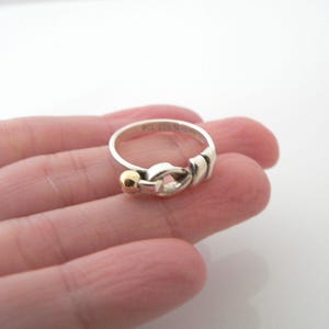 May include: A silver ring with a gold accent in the shape of a knot. The ring is on a person's finger.