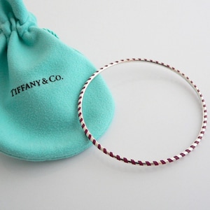 May include: A silver bangle bracelet with a white and burgundy striped design. The bracelet is shown with a Tiffany & Co. blue pouch.