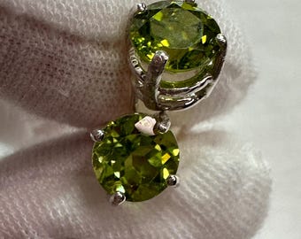 May include: A pair of stud earrings featuring round, faceted peridot gemstones set in silver-colored metal. The peridot stones have a vibrant, light green color and are held in place by four prongs. The earrings are displayed against a white background.