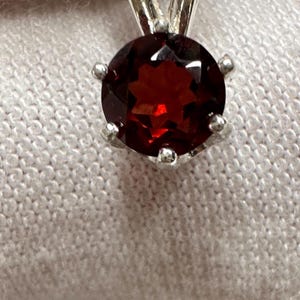 May include: A silver pendant featuring a round, faceted, deep red gemstone. The gemstone is set in a silver prong setting, and the pendant hangs from a silver bail. The gemstone has a star-like pattern in the center. Jewelry.