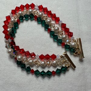 May include: A three-strand bracelet featuring red, green, and clear faceted beads, interspersed with faux pearls. The bracelet has a gold-tone clasp. The color scheme suggests a holiday theme.