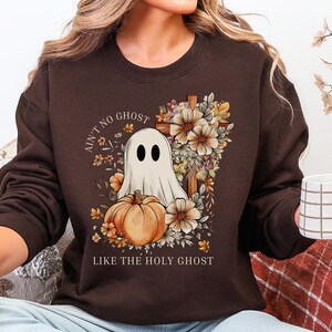 May include: A dark brown long-sleeve sweatshirt featuring a graphic of a white ghost, an orange pumpkin, and a wooden cross surrounded by autumn flowers. The text reads AIN'T NO GHOST LIKE THE HOLY GHOST in a circular font around the central design.