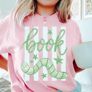 May include: Pale pink t-shirt with a green and white design. The design features the word "book" in cursive, a green worm, and green stars. The shirt is a light pink colour.