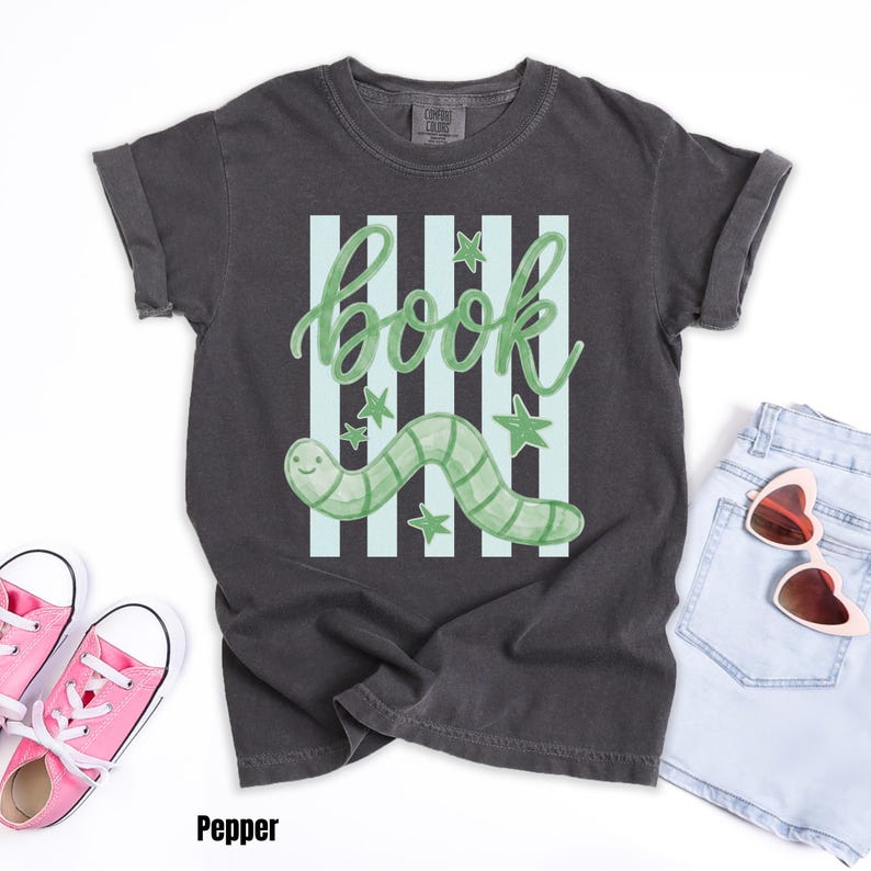 May include: Dark grey t-shirt featuring a green worm and the word "book". The design includes light blue stripes and stars. The shirt is styled with pink trainers and denim shorts.