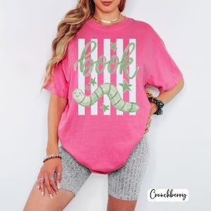 May include: A vibrant pink t-shirt with a playful design. The shirt displays the word "book" in green script, a green and white striped worm, and green stars. White vertical stripes are behind the text and worm.
