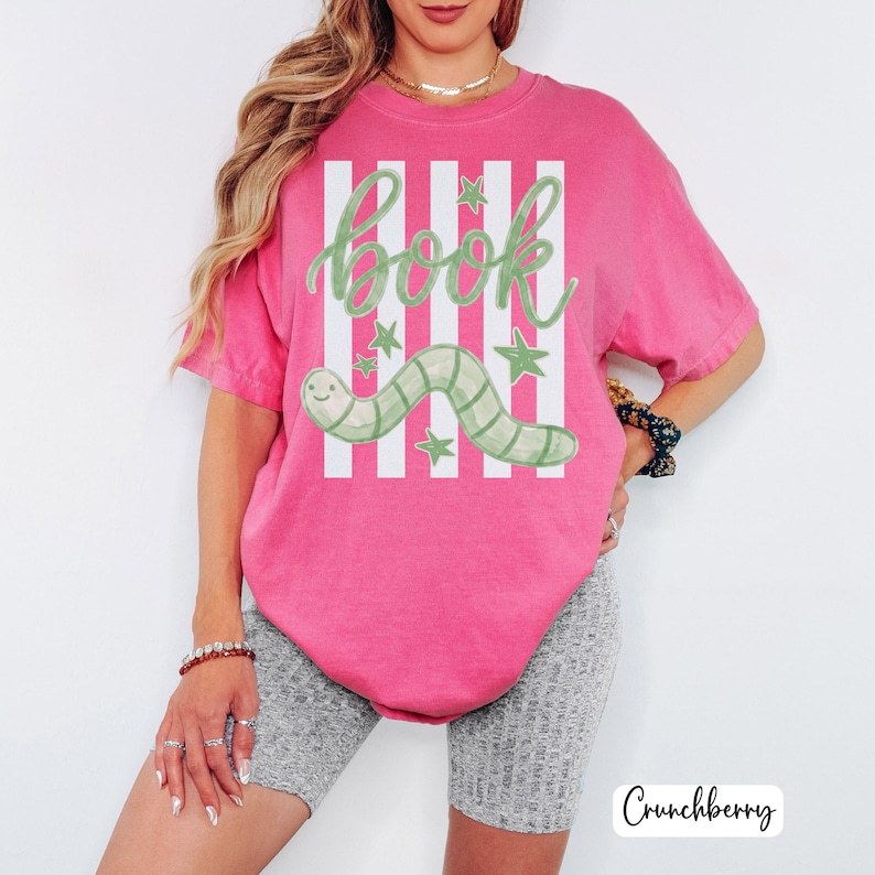 May include: A vibrant pink t-shirt with a playful design. The shirt displays the word "book" in green script, a green and white striped worm, and green stars. White vertical stripes are behind the text and worm.