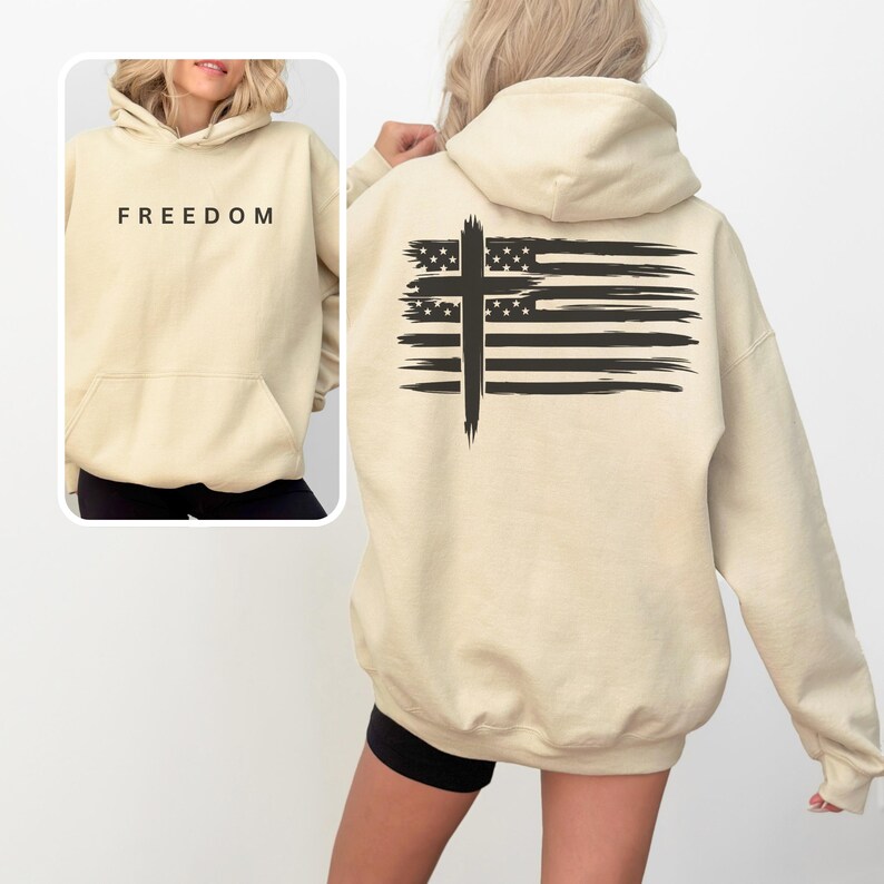 Op de afbeelding: Een beige hoodie met een voorzak en capuchon. Het woord "FREEDOM" is in het zwart op de voorkant gedrukt. De achterkant heeft een zwart kruisontwerp over een gestileerde Amerikaanse vlag.