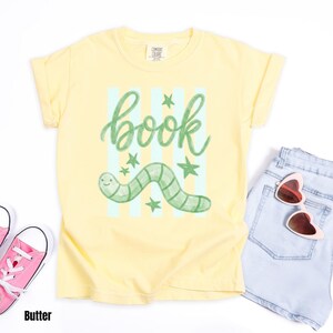 May include: A pale yellow t-shirt featuring a green worm illustration and the word "book". The design includes light blue vertical stripes and green stars. The shirt is styled with light blue denim shorts and pink sunglasses.