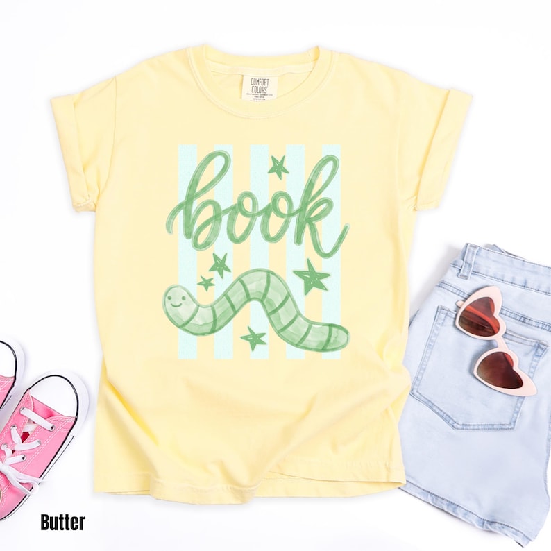 May include: A pale yellow t-shirt featuring a green worm illustration and the word "book". The design includes light blue vertical stripes and green stars. The shirt is styled with light blue denim shorts and pink sunglasses.