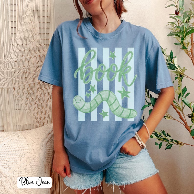 May include: A blue t-shirt featuring a green "book" design, a smiling worm, and stars. The graphic is set against light blue vertical stripes. The shirt is a relaxed, casual style.
