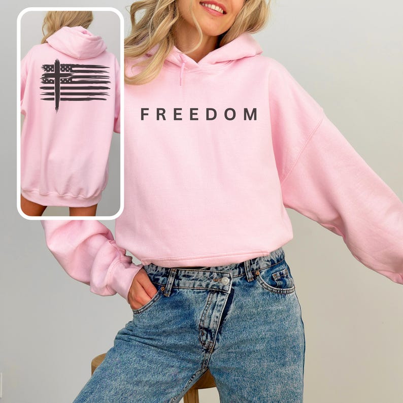 Op de afbeelding: Een lichtroze hoodie met het woord "FREEDOM" in zwarte letters op de borst gedrukt. De achterkant van de hoodie heeft een zwart kruis en een ontwerp van horizontale lijnen. De hoodie is gemaakt van zacht materiaal en heeft een capuchon.