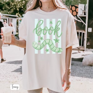 May include: Ivory-coloured t-shirt featuring a green worm graphic and the word "book" in a whimsical font. The design includes vertical stripes and stars. The shirt is a relaxed fit.