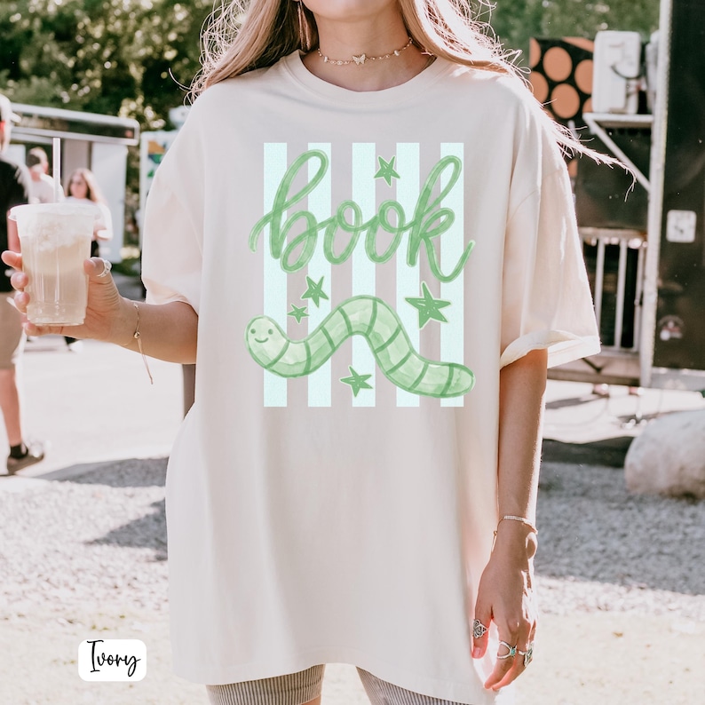 May include: Ivory-coloured t-shirt featuring a green worm graphic and the word "book" in a whimsical font. The design includes vertical stripes and stars. The shirt is a relaxed fit.