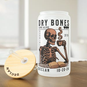 May include: White tumbler with a black and brown illustration of a skeleton holding a cup of coffee and a book. The text "DRY BONES COME ALIVE EZEKIEL 37:5 DECLAN 10-20-24" is printed on the tumbler.
