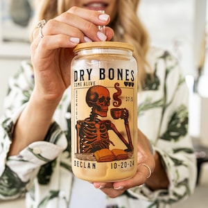 May include: A clear glass tumbler with a bamboo lid and a skeleton graphic. The graphic features a skeleton holding a cup of coffee and a book. The text on the tumbler reads "Dry Bones Come Alive Ezekiel 37:5 Declan 10-20-24".