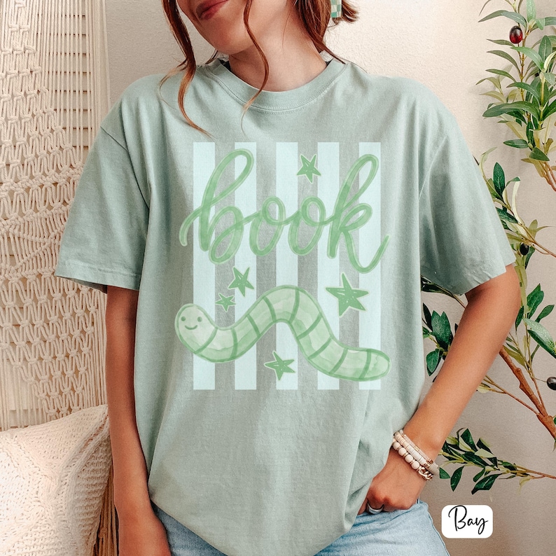 May include: Sage green t-shirt with a playful design. The design features the word "book" in a cursive font, a smiling green worm, and green stars. The background has vertical stripes in a lighter shade of green.