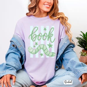 May include: A lilac t-shirt featuring the word "book" in green script, a green worm, and green stars. The design is set against light blue stripes. This is a casual wear item.