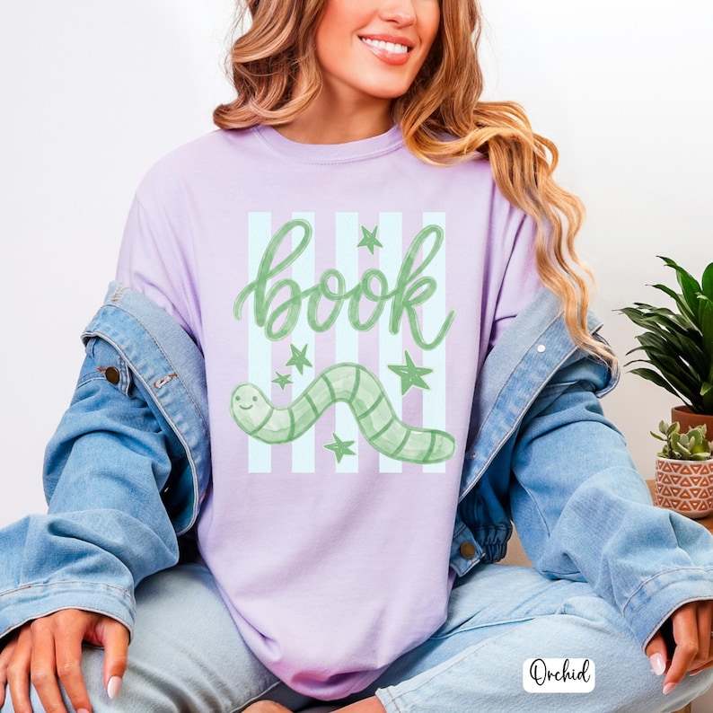 May include: A lilac t-shirt featuring the word "book" in green script, a green worm, and green stars. The design is set against light blue stripes. This is a casual wear item.