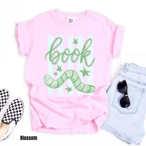 May include: A pale pink t-shirt featuring the word "book" in green script, a green worm, and green stars. The shirt is styled with light blue denim shorts, black sunglasses, and checkered slip-on shoes. The word "Blossom" is at the bottom.