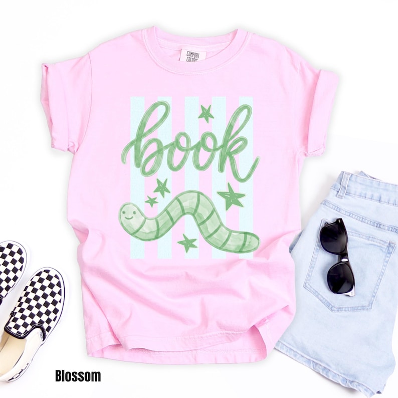 May include: A pale pink t-shirt featuring the word "book" in green script, a green worm, and green stars. The shirt is styled with light blue denim shorts, black sunglasses, and checkered slip-on shoes. The word "Blossom" is at the bottom.
