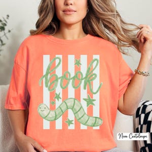 May include: Coral-coloured t-shirt with a whimsical design. The shirt features the word "book" in green script, set against white vertical stripes. A green worm and stars complete the design. The text "Neon Cantaloupe" is in the bottom right corner.