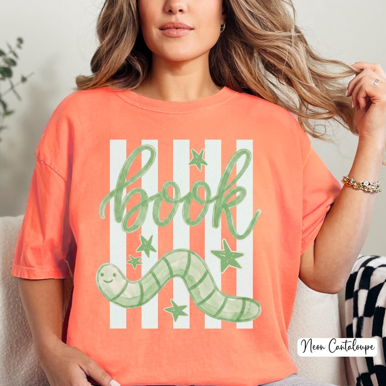 May include: Coral-coloured t-shirt with a whimsical design. The shirt features the word "book" in green script, set against white vertical stripes. A green worm and stars complete the design. The text "Neon Cantaloupe" is in the bottom right corner.