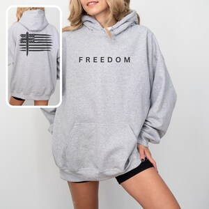 Op de afbeelding: Een lichtgrijze hoodie met het woord "FREEDOM" in zwart op de borst gedrukt. De achterkant van de hoodie heeft een zwart grafisch ontwerp dat lijkt op een vlag met een kruis. De hoodie heeft een capuchon en een voorzak.
