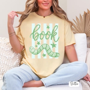 May include: Pale yellow t-shirt featuring the word "book" in green script, a green worm illustration, and green stars. The shirt has light blue and white vertical stripes behind the word "book" and worm.