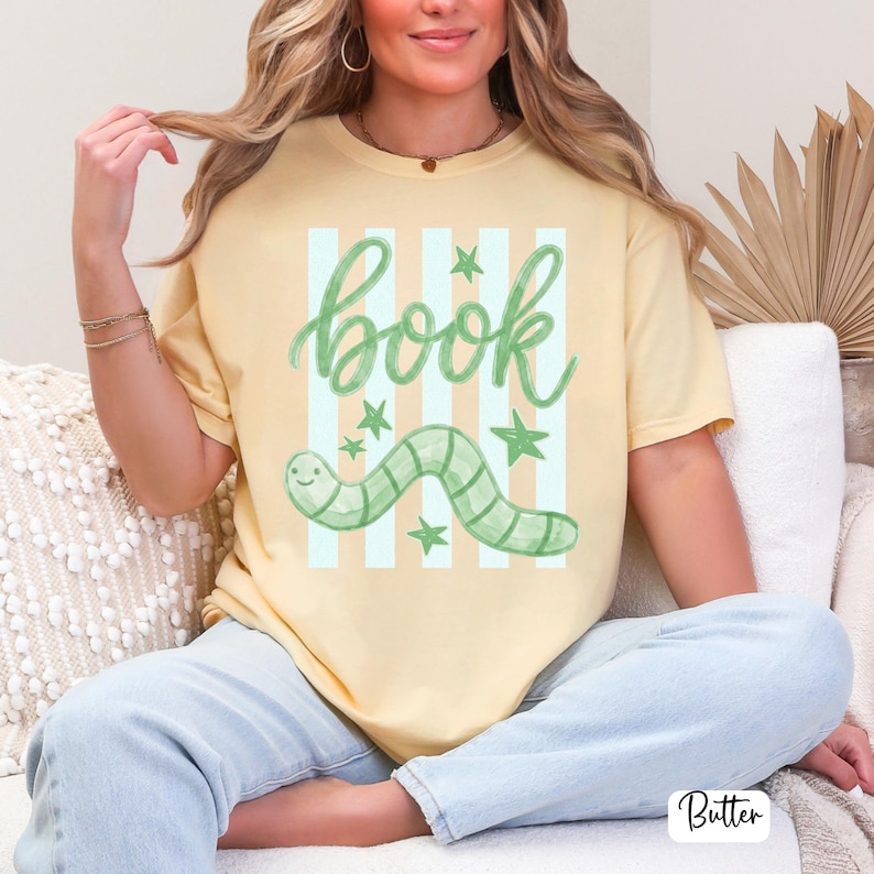 May include: Pale yellow t-shirt featuring the word "book" in green script, a green worm illustration, and green stars. The shirt has light blue and white vertical stripes behind the word "book" and worm.