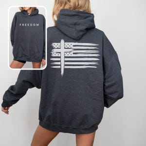 Op de afbeelding: Donkergrijze hoodie met een witte afbeelding van een kruis en een Amerikaanse vlag op de rug. De voorkant van de hoodie heeft het woord "FREEDOM" in het wit gedrukt. De hoodie heeft een capuchon en een voorzak.