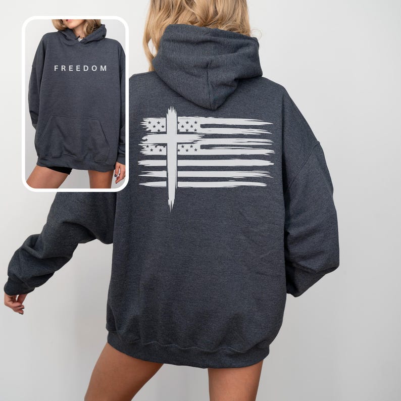 Op de afbeelding: Donkergrijze hoodie met een witte afbeelding van een kruis en een Amerikaanse vlag op de rug. De voorkant van de hoodie heeft het woord "FREEDOM" in het wit gedrukt. De hoodie heeft een capuchon en een voorzak.