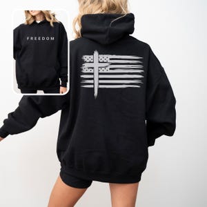 Op de afbeelding: Zwarte hoodie met het woord "FREEDOM" in het wit gedrukt op de voorkant. De achterkant heeft een verweerd Amerikaans vlagontwerp met een wit kruis eroverheen. De hoodie heeft een capuchon en een voorzak.