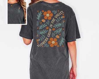 May include: Dark gray t-shirt with a floral design on the back. The design includes orange flowers, teal leaves, and the text "FLOWERS GROW IN THE VALLEY" in a curved font. A small floral design is on the front.