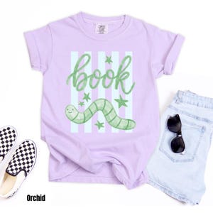 May include: Orchid-coloured t-shirt featuring a green worm illustration and the word "book" in cursive. The design includes green stars and vertical stripes. The shirt is paired with light blue denim shorts and black and white checkered slip-on shoes.