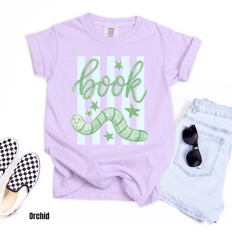 May include: Orchid-coloured t-shirt featuring a green worm illustration and the word "book" in cursive. The design includes green stars and vertical stripes. The shirt is paired with light blue denim shorts and black and white checkered slip-on shoes.