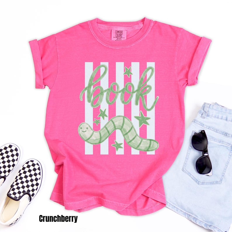 May include: A vibrant pink t-shirt with a green and white design. The design includes the word "book", a smiling green worm, and stars. The shirt is styled with denim shorts, sunglasses, and black and white checkered slip-on shoes.