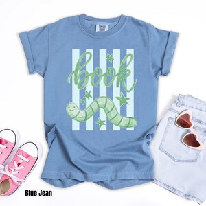 May include: A blue t-shirt featuring a green worm design and the word "book". The illustration includes green stars and vertical stripes. The shirt is styled with pink trainers and light blue denim shorts.