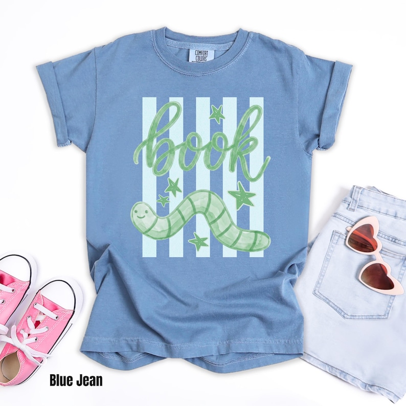 May include: A blue t-shirt featuring a green worm design and the word "book". The illustration includes green stars and vertical stripes. The shirt is styled with pink trainers and light blue denim shorts.