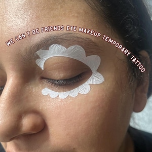 May include: Close-up of a person's eye with a white, scalloped eye makeup stencil applied. The text "WE CAN'T BE FRIENDS EYE MAKEUP TEMPORARY TATTOO" is arched above the eye. The skin tone is light with a slight pink hue.