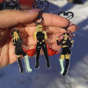 May include: Three acrylic keychains featuring stylized illustrations of women in fashionable outfits. Each keychain has a heart-shaped charm and a silver-colored heart-shaped clasp. The women are depicted in black clothing with colorful accents.