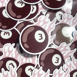 May include: A selection of stickers depicting a maroon 8-ball with a white number 3, set against pink and white flames. The stickers have a white border and are arranged on a light pink surface.