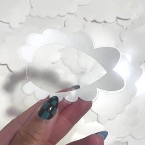 Könnte beinhalten: Eine Sammlung weißer, wolkenförmiger Aufkleber mit gewelltem Rand. Ein Aufkleber wird hochgehalten und zeigt einen klaren, tropfenförmigen Ausschnitt in der Mitte. Der Hintergrund ist ein zartes Rosa, mit weiteren Wolkenaufklebern verstreut.