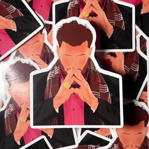 May include: A collection of stickers featuring a stylized illustration of a person in a black suit, pink shirt, and a patterned scarf. The person has their hands clasped in front of their face, wearing a gold ring. The stickers are on a white background.